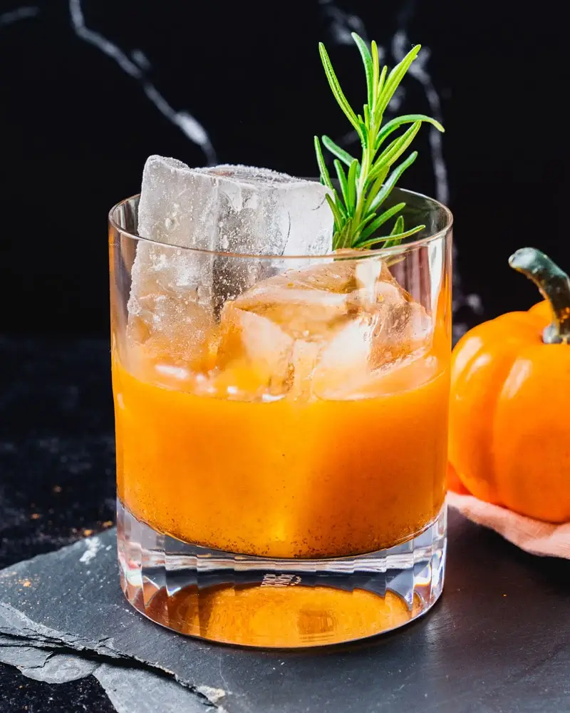 pumpkin old fashioned in a glass with ice and a sprig of rosemary set against a dark background