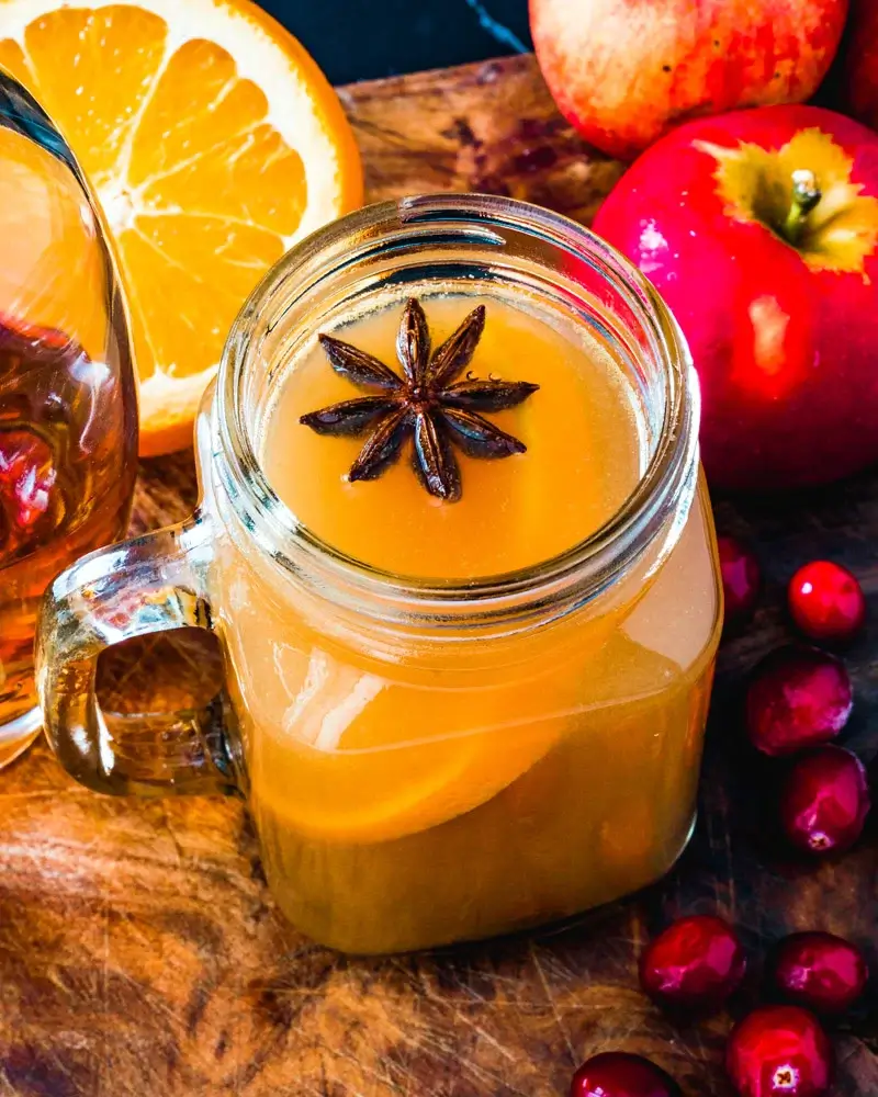 hot spiked apple cider in a glass jar with a background of oranges and apples