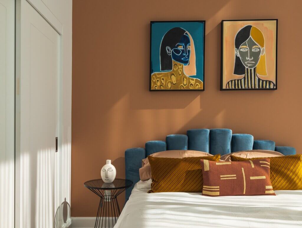 Bedroom with a dark peach accent wall, blue and yellow artwork, and a bed with a blue headboard, gold and orange pillows, and a white beadspread.