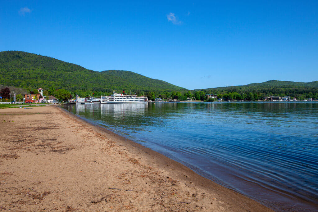View of beach in Lake George with sand on the left, the lake on the right, and mountains and a cruise boat in the background