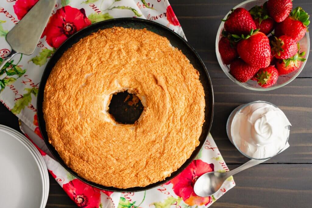Angel food cake in a pan next to fresh strawberries and whipped cream