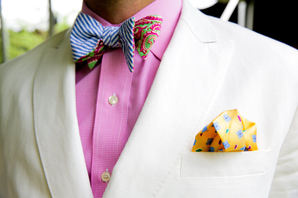 close up of a man in a white linen suit jacket with a pink shirt and colorful bowties and pocket square